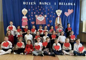Wnuczkowie klasy 1a na tle dekoracji siedzą i trzymają w dłoniach serduszka dla swoich dziadków i babć.