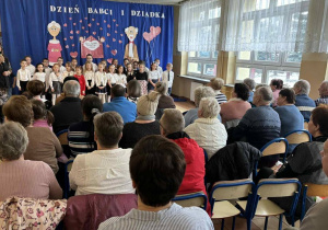 Babcie i dziadkowie siedzą na krzesłach zasłuchani w występy swoich wnucząt.