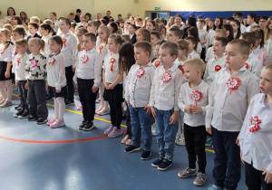 Duża grupa uczniów ubrana na galowo stoi śpiewając hymn.