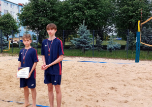 Natan i Piotr z dyplomem i medalmi na szyi stoją na boisku do piłki siatkowej plażowej
