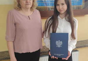 Ula z panią Elżbietą Kafar na tle sztandaru szkoły. Ula w dłoni trzyma dyplom za udział w finale wojewódzkim z języka polskiego