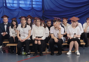Duża grupa uczniów klasy 8b w biretach na głowie siedzi na ławeczkach podczas zakończenia klas ósmych.