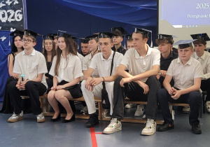 Duża grupa uczniów klasy 8a w biretach na głowie siedzi na ławeczkach podczas zakończenia klas ósmych.