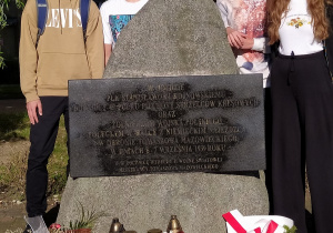 Kamień z zamontowana płyta poświęcony pamięci Hojnowskiego. Przed pomnikiem leżą białe i czerwone kwiaty. Za pomnikiem stoją dwie dziewczyny i dwóch chłopaków. W tle drzewa.