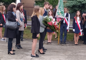 Rzędy osób ustawione do składania kwiatów przed pomnikiem. Za nimi szereg składający się z pocztów sztandarowych. w tle drzewa i kamienica.