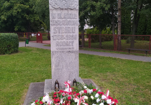 Pomnik Sybiraków. Wysoki obelisk z wyrzeźbionym orłem. Pod obeliskiem leżą wiązanki z białych i czerwonych kwiatów. W tle drzewa.