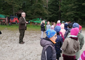 Grupa dzieci stoi po prawej stronie. Przed nimi stoi leśniczy .W tle maszyny rolnicze i las.