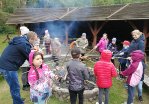 Ognisko otoczone kamieniami. Wokół niego dzieci z patykami na których nadziane są kiełbaski. Po lewej stronie ogniska mężczyzna pochyla się z patykiem a po prawej stoi uśmiechnięta mama. W tle drewniana wiata do biesiadowania.