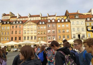 Grupa uczniów na tle kamieniczek Starego Miasta rożświetlonych słońcem.