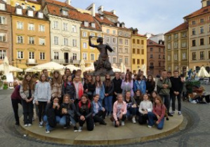 Grupowe zdjęcie uczniów na tle pomnika Małej Syrenki.W tle rozświetlone słońcem kamieniczki Starego Miasta.