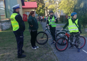 Wejście do miasteczka ruchu drogowego. Na pierwszym planie stoi policjant w odblaskowej kamizelce, nauczycielka oraz dwóch uczniów z rowerami w kaskach i kamizelkach. W tle budynki mieszkalne.