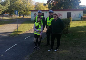 Miasteczko ruchu drogowego. Na pierwszym planie stoją policjanci z drogówki, nauczycielka i dwie uczennice w kaskach i kamizelkach odblaskowych. W tle drzewa, budynek techniczny i teren zielony.