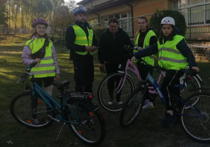 Miasteczko ruchu drogowego. Na pierwszym planie stoją policjant z drogówki, nauczycielka i trzy uczennice w kaskach i kamizelkach odblaskowych ze swoimi rowerami. W tle drzewa, budynek i teren zielony.