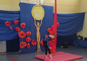 Sala gimnastyczna. Dziewczynka trzyma czerwoną szarfę i kłania się widowni. i chłopiec w środku stoją z mikrofonami w dłoniach. W tle niebieska dekoracja z czerwonymi różam