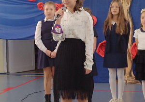 Sala gimnastyczna. Dziewczynka ubrana na galowo z mikrofonami w dłoniach śpiewa . W tle chórek składający się z kilku dziewczyn stojący na niebieskim tle.