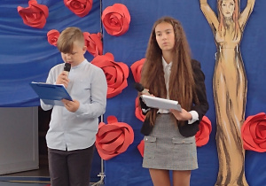 Sala gimnastyczna. Chłopiec i dziewczyna ubrani na galowo z mikrofonami w dłoniach. W tle niebieska dekoracja z czerwonymi różami.