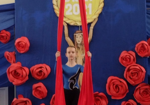 Sala gimnastyczna. na czerwonym materacu sroi dziewczyna i trzyma w dłoniach dwie czerwone szarfy.W tle niebieska dekoracja z czerwonymi różami.