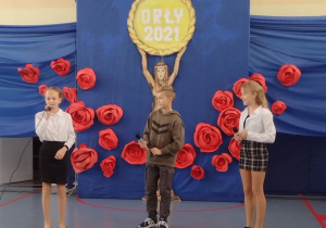 Sala gimnastyczna. Dwie dziewczynki i jeden chłopiec w środku trzymają mikrofony. W tle niebieska dekoracja z czerwonymi różami
