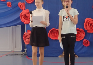 Sala gimnastyczna. Dwie dziewczynki ubrane na galowo z mikrofonami w dłoniach. W tle niebieska dekoracja z czerwonymi różami.