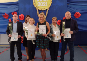 Sala gimnastyczna. Cztery nauczycieli i dwóch nauczycieli stoi z dyplomami w rękach na tle niebieskiej dekoracji z czerwonymi różami.