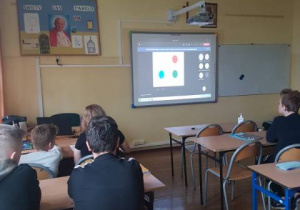Wnętrze klasy. Uczniowie słuchają wykładu on-line. W tle widać obraz wyświetlony na tablicy multimedialnej.