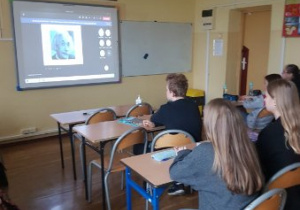 Wnętrze klasy. Uczniowie słuchają wykładu on-line. W tle widać obraz wyświetlony na tablicy multimedialnej.