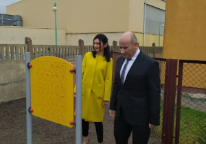 Pan Prezydent w garniturze i pani Beata Stańczyk stoją przed tablicą na placu zabaw. W tle budynki szkoły.