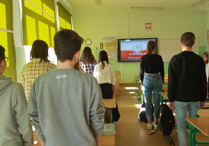 Uczniowie zwróceni przodem do tablicy interaktywnej śpiewają hymn Polski. W tle tablica z wyświetlonym tekstem.