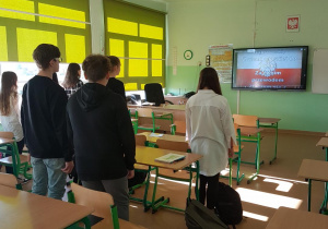 Uczniowie zwróceni przodem do tablicy interaktywnej śpiewają hymn Polski. W tle tablica z wyświetlonym tekstem.