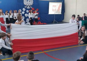 Uczniowie ubrani na galowo trzymają w dłoniach rozciągniętą w poziomie flagę Polski. W tle grupa uczniów.
