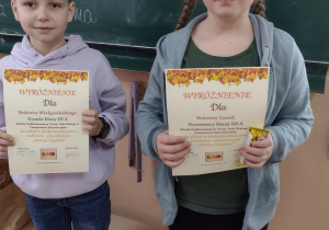 Chłopiec i dziewczynka stoją przed zielona tablicą. W dłoniach trzymają kolorowe dyplomy.