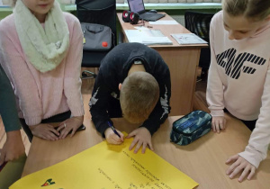 Dwie dziewczynki stoją przy stoliku. Chłopiec podpisuje leżący na stoliku dekalog. W tle biurko.