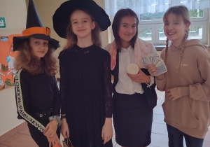 Cztery dziewczynki stoją obok siebie. Dwie z nich są ubrane na czarno i maja kapelusze czarownic . dziewczynka z prawej stronie trzyma w dłoni karty. w tle okno.