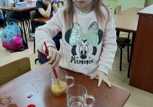 Dziewczynka w jasnej bluzce miesza żóltą wodę w szklance. Obok na stoliku stoi druga szklanka z wodą i czerwona farba. W tle dzieci siedzą w ławkach. stoliku stoi