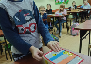 Chłopiec stoi przed stolikiem.Na stolikiem stoi pudełko z którego w różnych miejscach wystają różne sznurki zakończone metalowymi kółkami. W tle dzieci siedzą w ławkach.