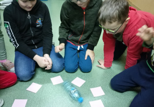 NA podłodze leżą ułożone w krąg białe karteczki. W środku kręgu lezy plastikowa butelka. Wokół karteczek siedzą chłopcy.