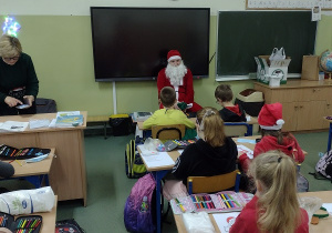 Uczniowie siedzący w ławkach na przeciwko Świętego Mikołaja. W tle zielona tablica.