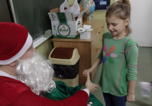 Święty Mikołaj podaje rękę dziewczynce w zielonej sukience. w tle zielona tablica i plakat z układem słonecznym.