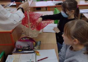 Święty Mikołaj z koszykiem cukierków częstuje dzieci siedzące w ławkach.