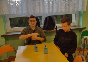 Dwóch chłopców siedzi przy żółtym stole. Na stole stoi woda do picia.