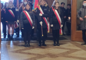 Dwie dziewczyny i chłopiec z biało czerwonymi szarfami na pierwszym planie. Chłopiec trzyma sztandar szkoły .W tle inne poczty sztandarowe.