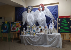Dwa olbrzymie anioły przed którymi stoi stół z aniołkami wykonanymi z różnych materiałów. Po prawej i lewej stronie aniołów stoją sztalugi z pracami plastycznym.