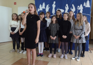 Dziewczynka w czarnej sukience z mikrofonem w dłoni. W tle stoi chórek składający się z kilkunastu dziewczynek ubranych galowo .W tle biało niebieska dekoracja .