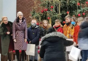 Kobieta stojąca tyłem robi zdjęcie grupie dzieci i dwóm kobietom. Dzieci stoja na tle wielkiej choinki.