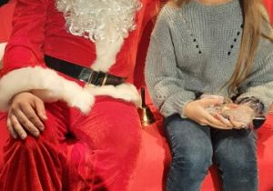Dziewczynka w maseczce siedzi na czerwonej kanapie w towarzystwie Świętego Mikołaja.