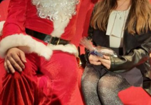 Dziewczynka z długimi włosami w maseczce siedzi obok Świętego Mikołaja.