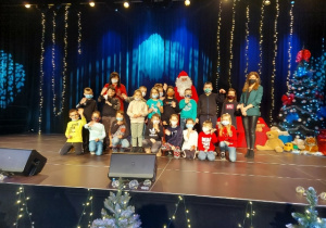 Grupa uczniów w towarzystwie Świętego Mikołaja i nauczycielek. W tle scena nastrojowo oświetlona światełkami.