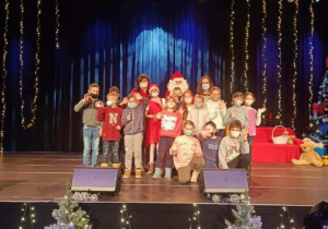 Grupa dzieci w towarzystwie Świętego Mikołaja na scenie . Z przodu sceny choinki oprószone śniegiem.