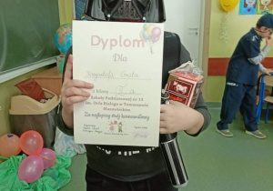 Chłopiec przebrany za lorda Vadera trzymający dyplom. W tle wnętrze klasy.