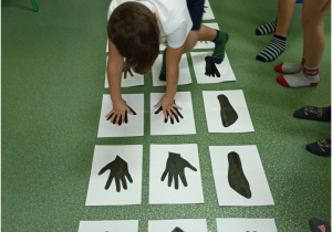 Na podłodze w trzech rzędach po rozłożono znaki graficzne dłoni i stóp . Chłopiec na stopami dotyka plansz z e znakiem stóp a dłońmi plansz ze znakami dłoni. w tle stopy chłopców w kolorowych skarpetkach.